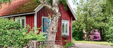 Una casa in legno rossa con un muro in pietra, un albero di fronte e un giardino con fiori rosa.