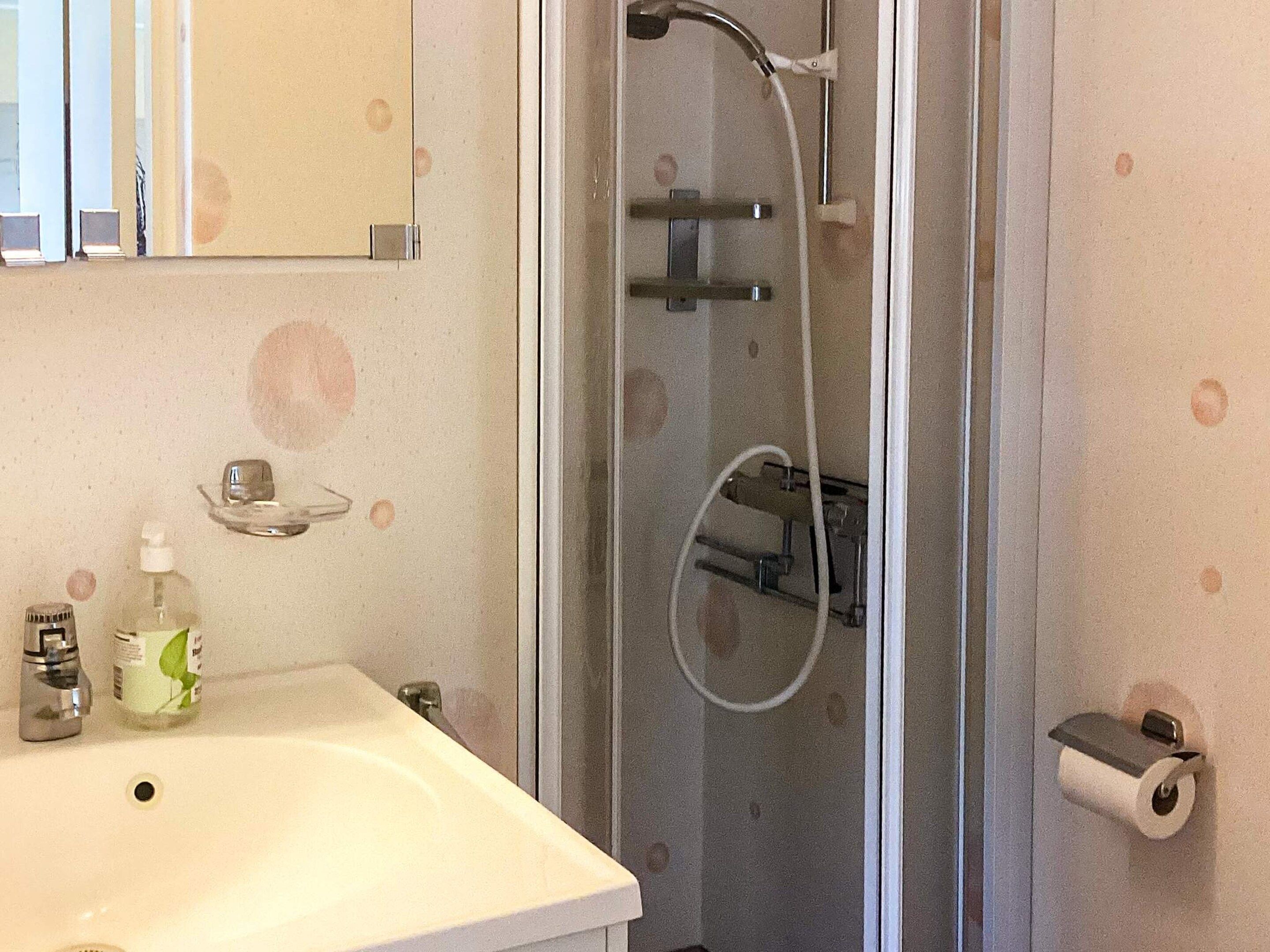 Un baño con lavamanos, espejo, ducha con puerta de vidrio y soporte para papel higiénico.
