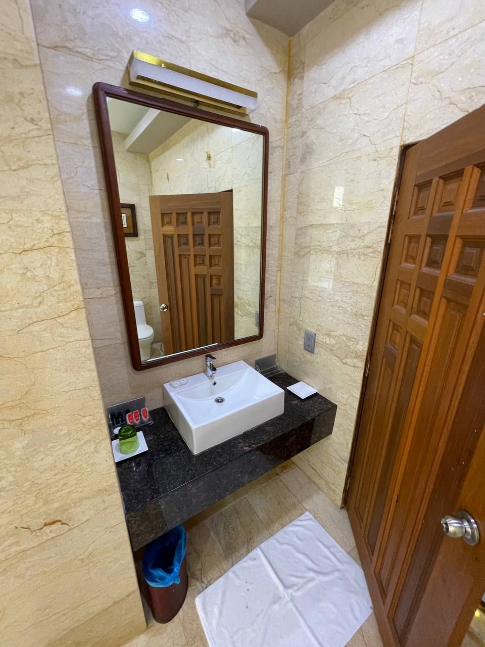 Une salle de bain avec un mur en marbre, un grand miroir, un lavabo blanc, une porte en bois et une serviette sur le sol.