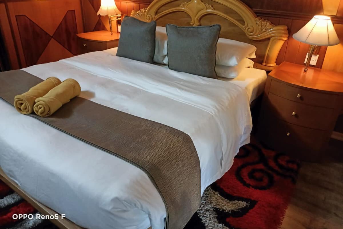 Un lit bien fait, avec une couette blanche, deux oreillers et une serviette pliée. On y trouve deux tables de chevet avec des lampes, une tête de lit en bois et un tapis rouge à motifs.