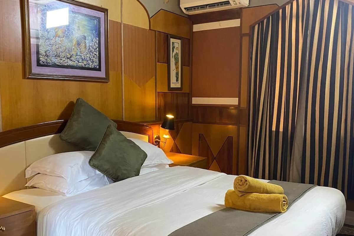 Une chambre d’hôtel avec un lit, des oreillers, une table de chevet, un appareil de climatisation, un tableau encadré au mur et un rideau à rayures.