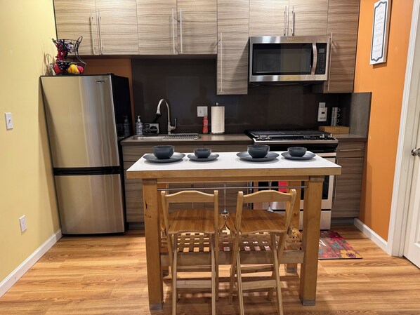 A compact kitchen with a microwave, stove, and refrigerator. There is a wooden table with four bowls and two chairs.