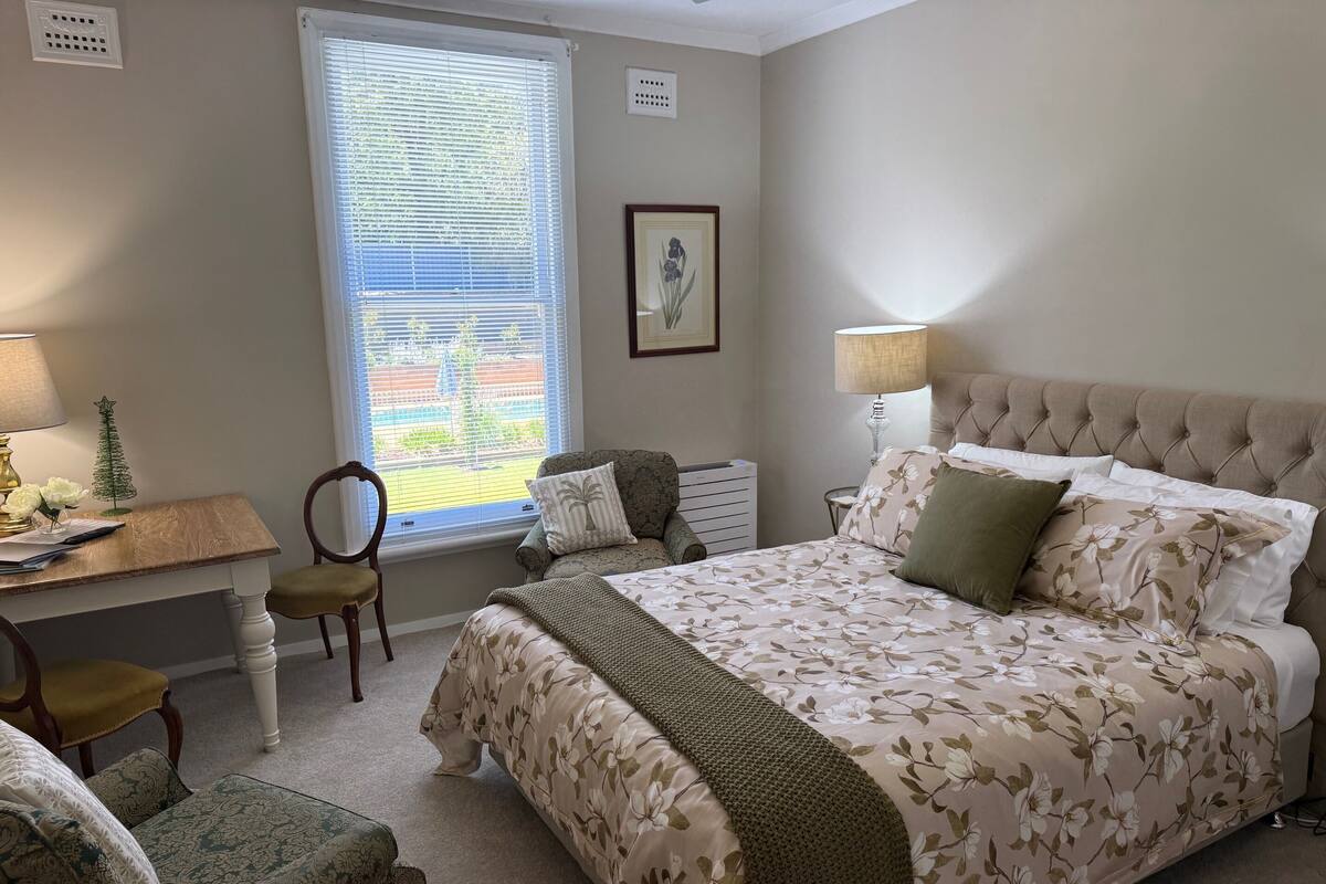A bedroom with a bed, bedside table, lamp, chair, and a window with blinds.