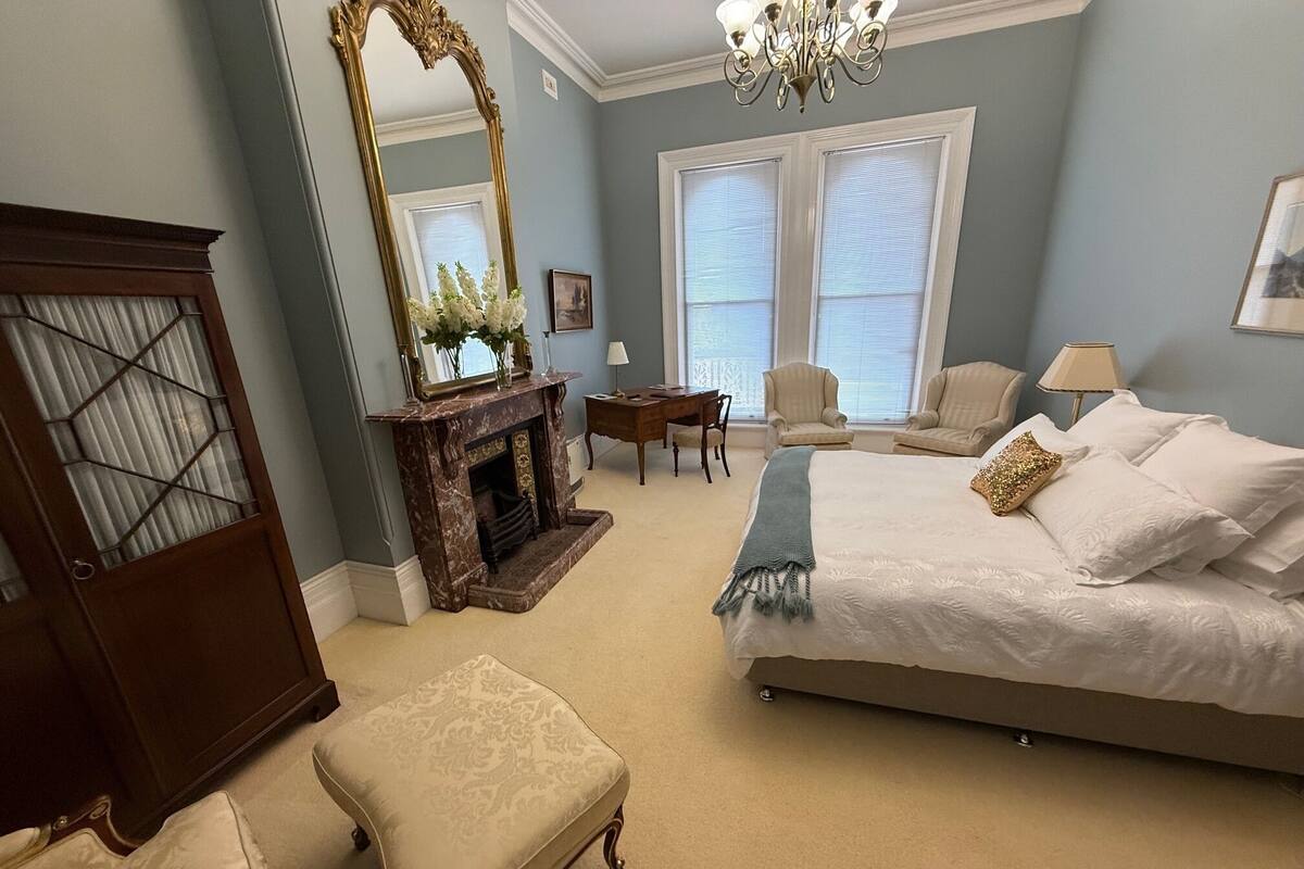 A bedroom with a large bed, a fireplace, a mirror, and two armchairs.