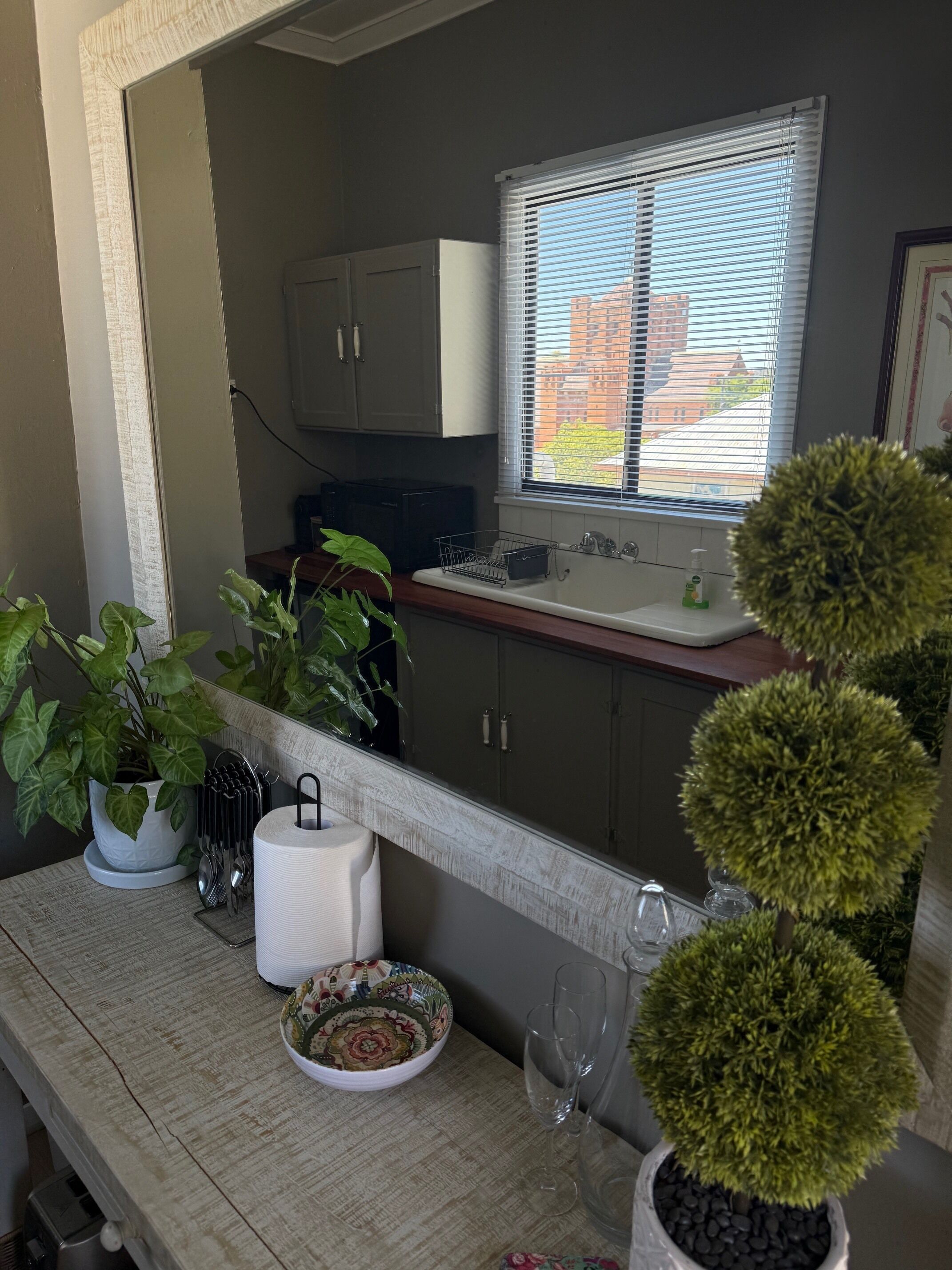 A kitchen with cabinets, a window with blinds, a sink, a potted plant, a dish rack, a bowl, and a wine glass.