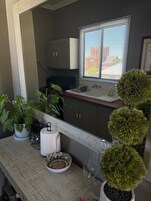 Una cocina con armarios, una ventana con persianas, un lavamanos, una planta en maceta, un escurreplatos, un bol y una copa de vino.