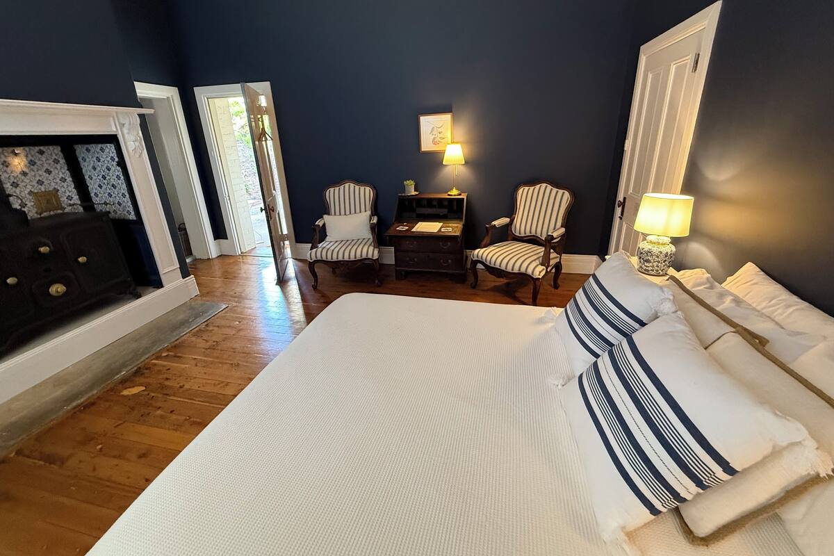 A bedroom with a large bed, two chairs, a desk, a lamp, and a fireplace.