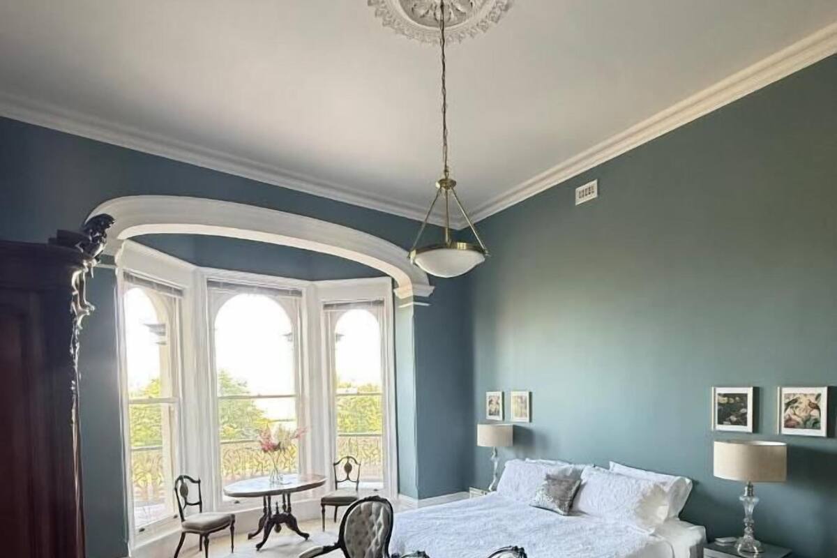 A bedroom with a large bed, a sitting area with a table and chairs, and a view of the outdoors through large windows.