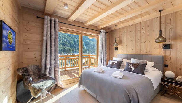 A wooden cabin bedroom with a bed, a fur chair, and a balcony with a view of the forest. - 1/2 Chalet: Alpine sweetness at Linga (CHATEL)