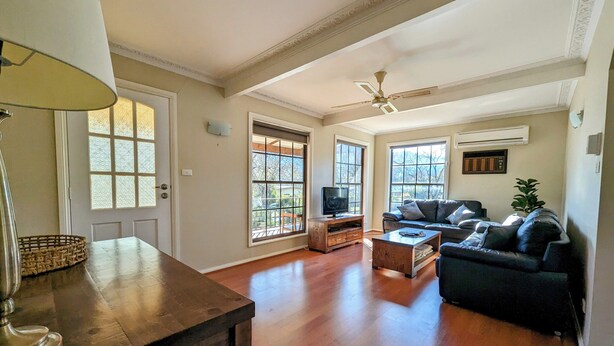A spacious living room with hardwood floors, a ceiling fan, a television on a wooden stand, a dark-colored sofa, and a potted plant. - Cupie’s Cabin (Talbingo)