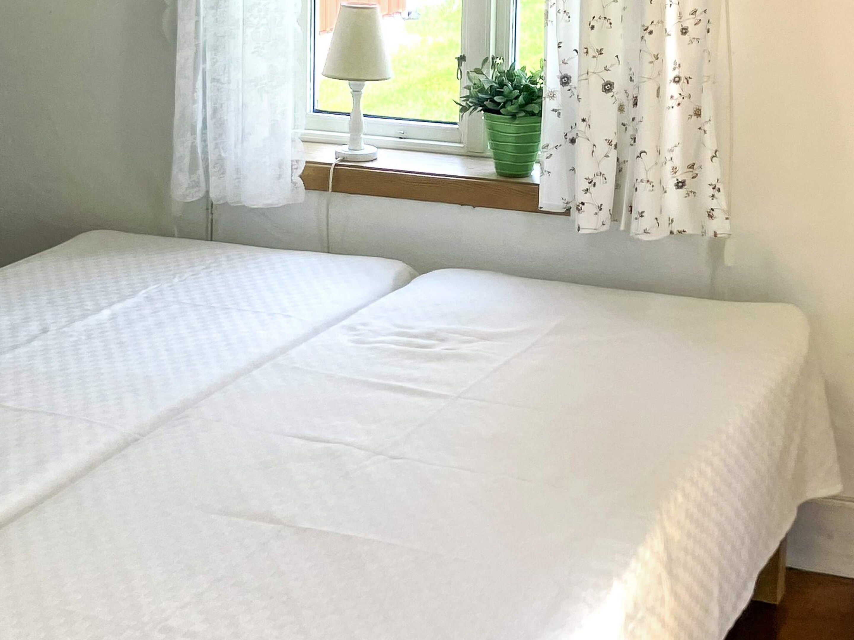 A room with two beds, a window with curtains, a lamp, and a plant on a windowsill.