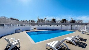 Une piscine rectangulaire à l’eau d’un bleu limpide, entourée d’une terrasse en béton. On y trouve des chaises longues blanches, dont les dossiers sont tournés vers la piscine, et une clôture blanche qui délimite l’espace.