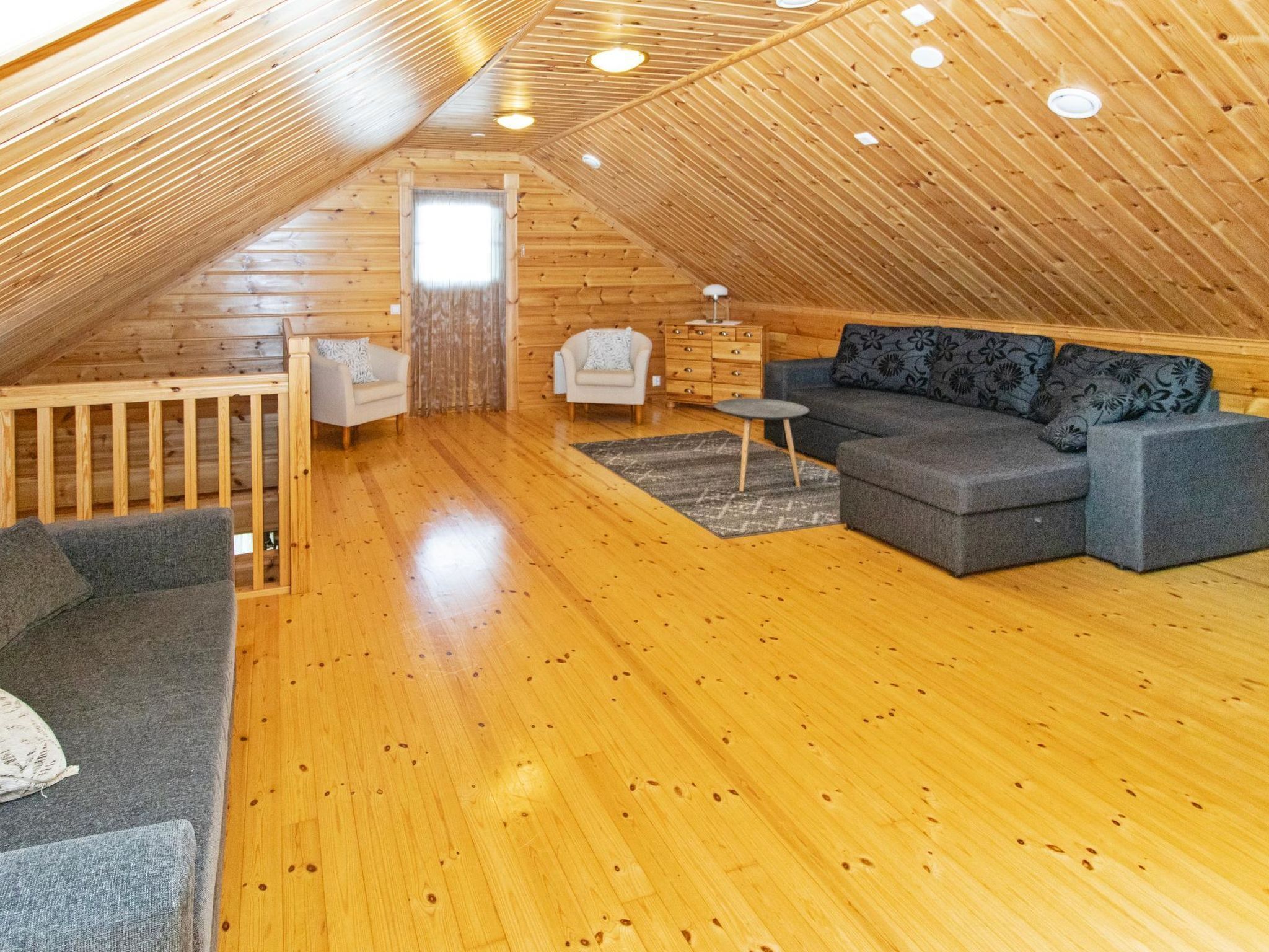 Acogedor dormitorio en el ático con paredes de madera y techo inclinado, amoblado con un sofá gris, una mesita redonda y una silla blanca.