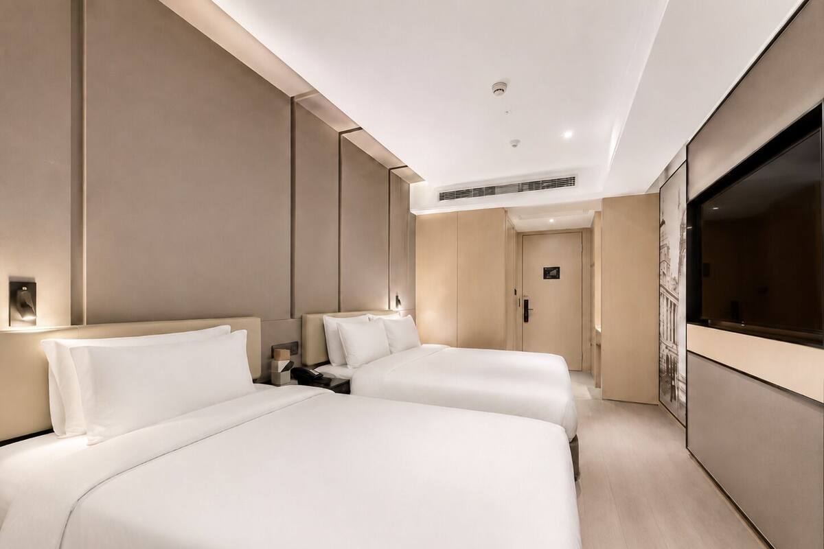 A hotel room with two beds, a TV, a door, and a wall-mounted light fixture.