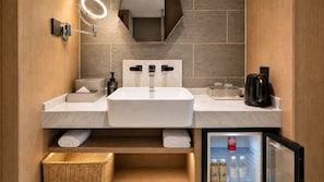 Une salle de bain moderne dotée d’un double lavabo, d’un miroir avec éclairage intégré, d’une machine à café et d’un mini-réfrigérateur contenant des bouteilles d’eau.