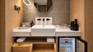 Un baño moderno con lavamanos doble, espejo con iluminación integrada, cafetera y mini refrigerador con agua embotellada.