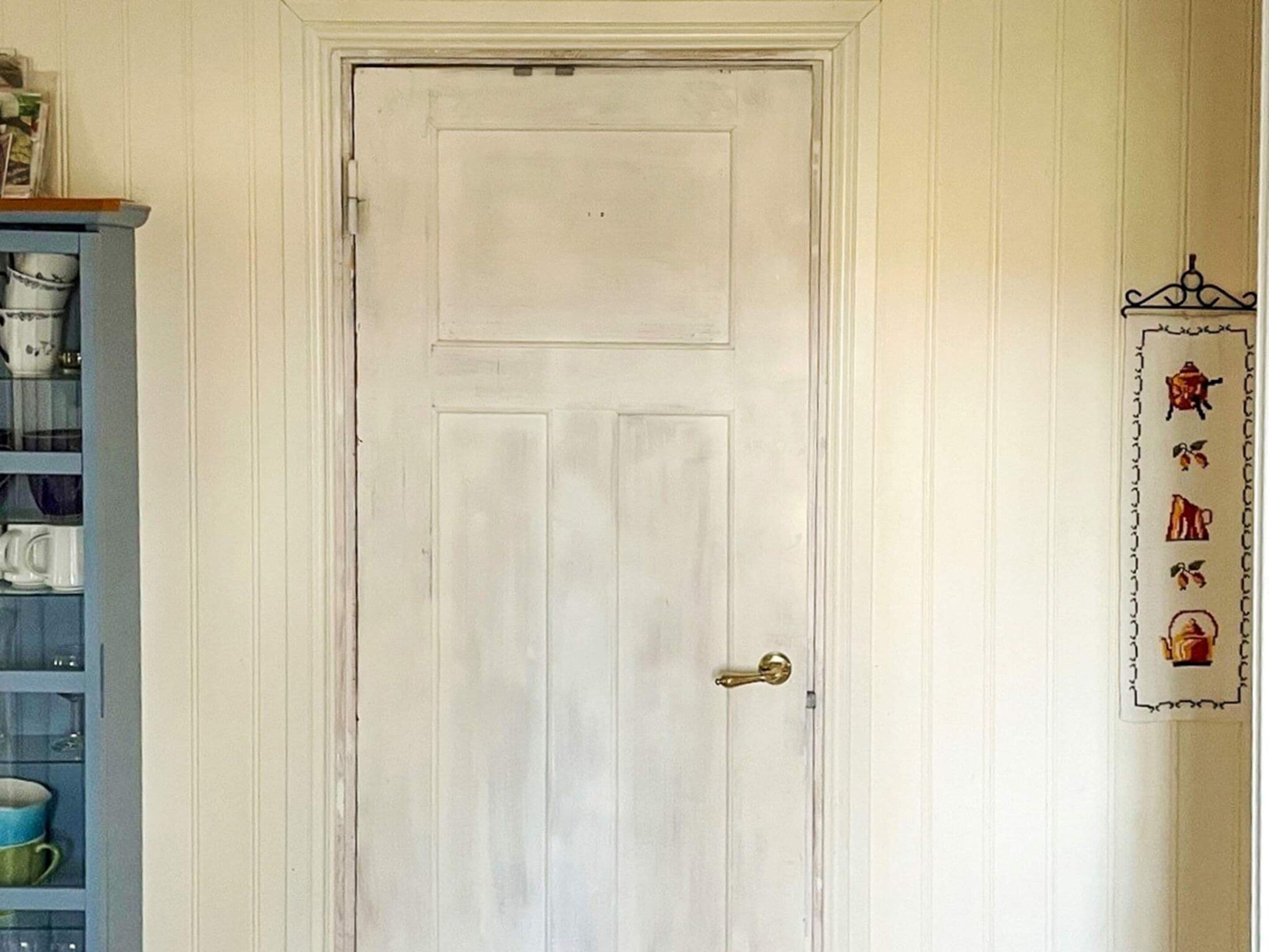 Una puerta blanca con pomo de latón, un estante con utensilios de cocina a la izquierda y una decoración montada en la pared a la derecha.