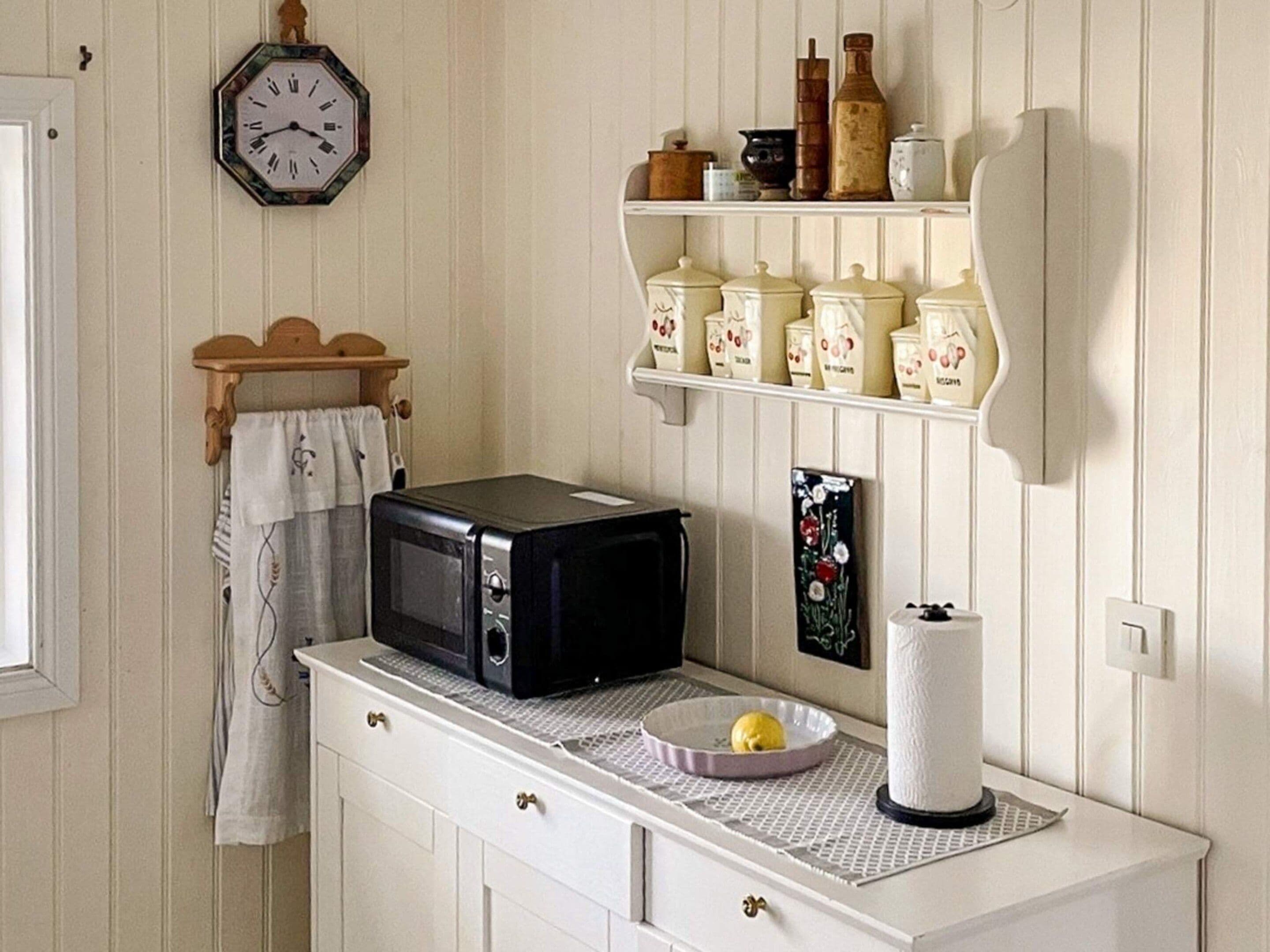 Una cocina con un microondas en la encimera, un reloj en la pared y estantes con frascos y recipientes.