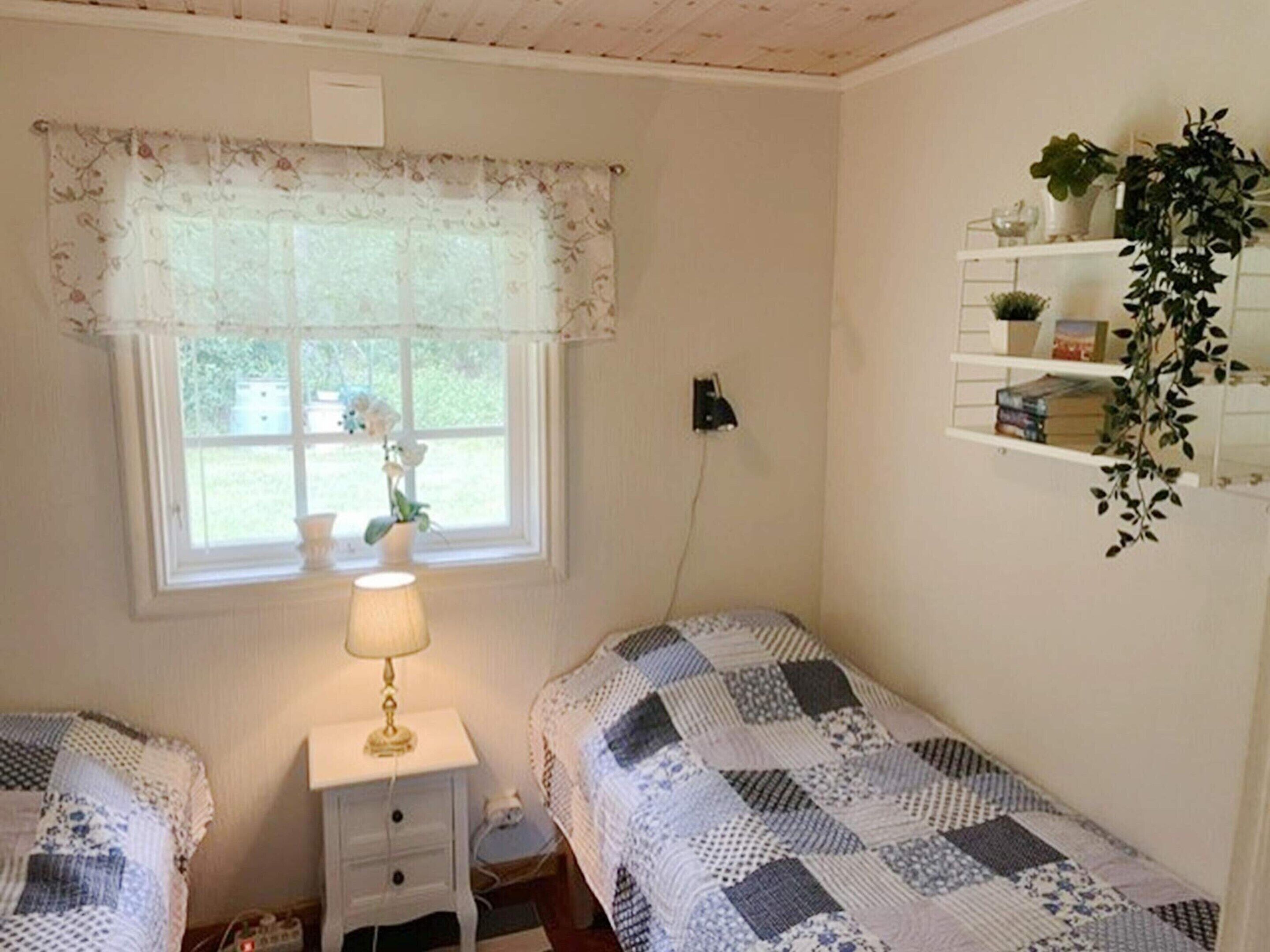 Une chambre avec deux lits, une fenêtre ornée de rideaux à motifs floraux, une lampe de chevet et une étagère avec des plantes.