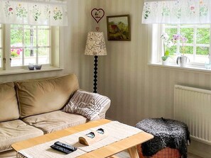 Una habitación acogedora con un sofá color beige, una mesa de centro de madera con un control remoto, una lámpara estampada y una ventana con vistas a zonas verdes.