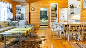 A cozy cabin interior with a wooden ceiling, a dining area with a table and chairs, a sofa with cushions, a large rug, and a staircase leading to another room.