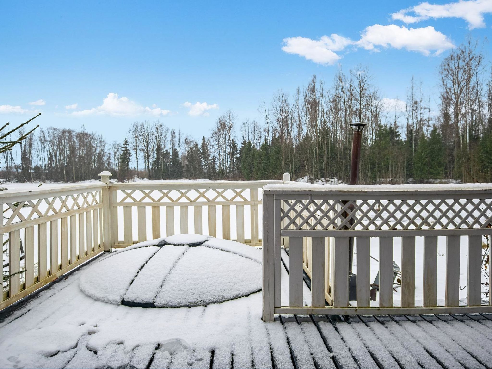 A snow-covered deck with a lattice railing, surrounded by a snowy landscape and trees.