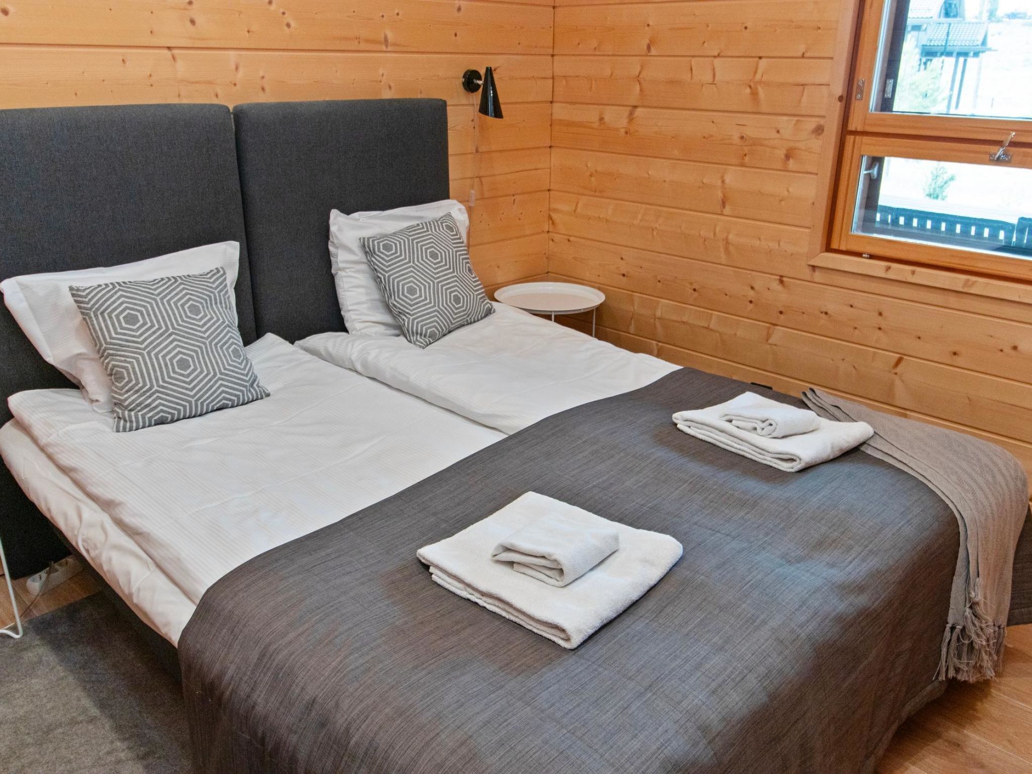 Ein Doppelbett mit grauer Bettwäsche und zwei Kissen, ein kleiner runder Tisch und ein Fenster mit Blick auf Gebäude.