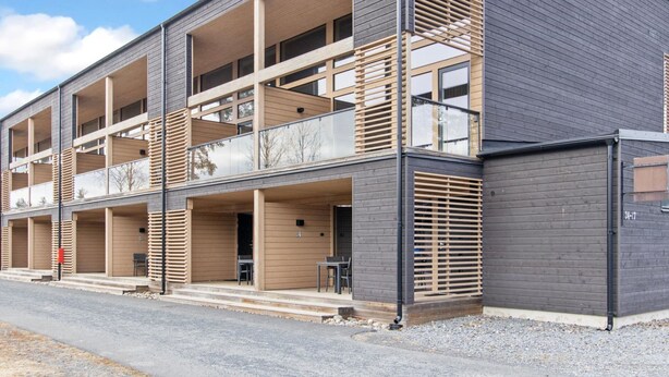 A modern multi-story building with balconies, large windows, and a gravel driveway.
