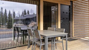 A modern outdoor dining area with a wooden deck, a glass door leading to a view of trees and a building, and a black door with a metal grille.
