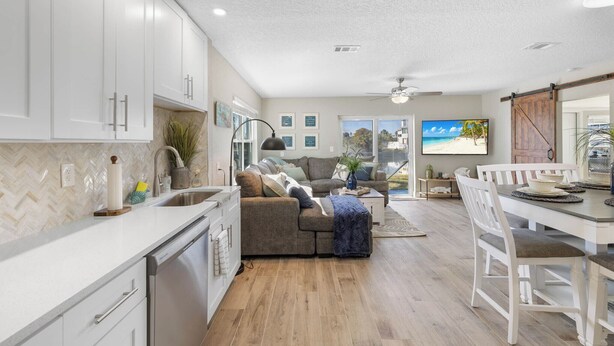 A modern kitchen with white cabinets, a dining area with a white table and chairs, and a living room with a gray sofa and a TV displaying a beach scene. - Boat, Kayak & Relax: Stunning Waterfront Home w/ Sunsets and Direct Gulf Access (Hernando Beach)