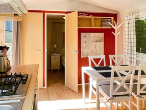Une pièce comprenant une table à manger et des chaises, un coin cuisine avec un fourneau, un mur rouge orné d’un tableau encadré et une porte-fenêtre donnant sur une autre pièce.