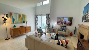 A spacious living room with a sofa, a coffee table, a TV, a fireplace, and a large window with curtains. - Downtown Palm Springs Holiday Loft, View Patio (Palm Springs)