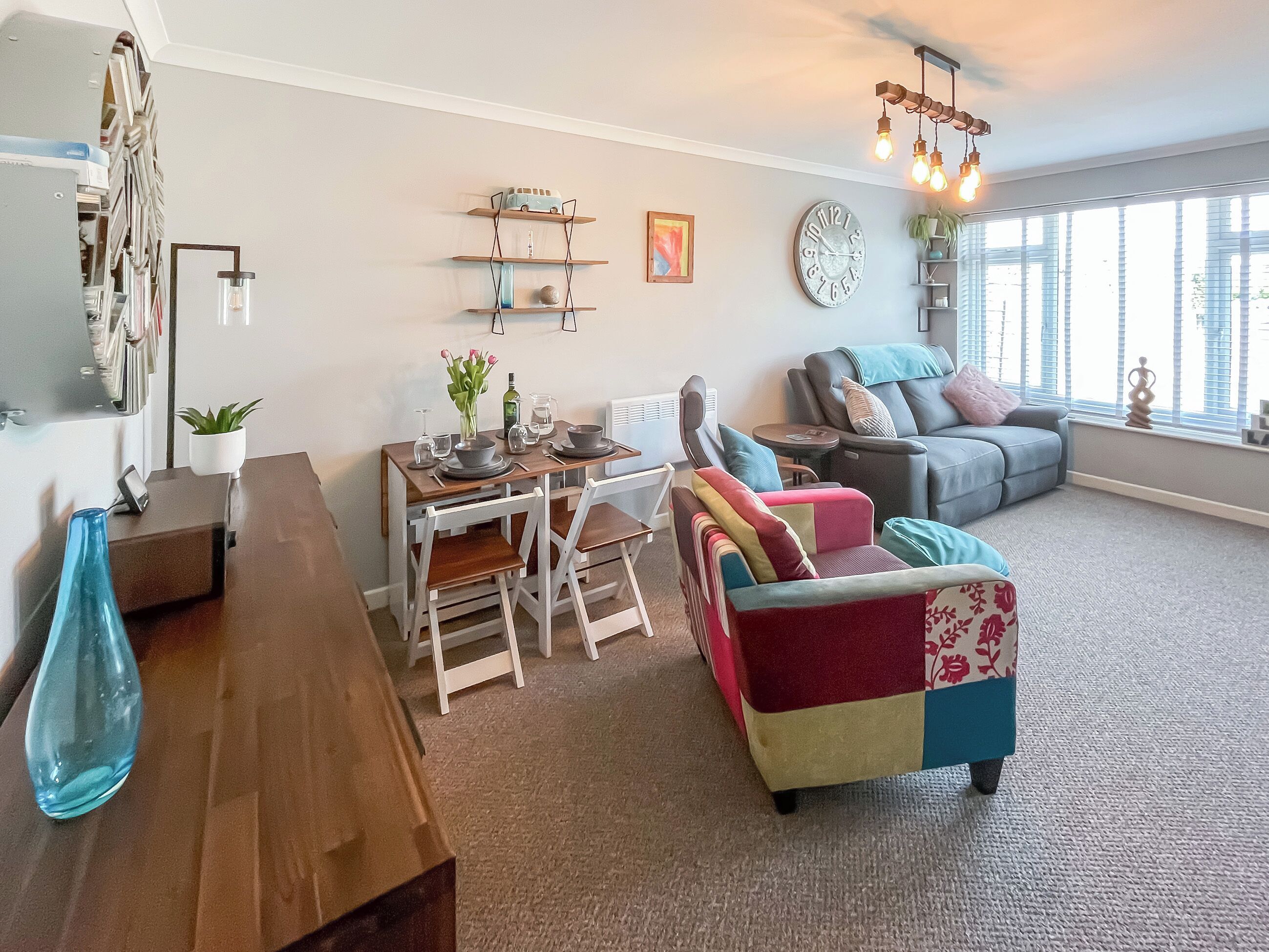 A living room with a dining table, chairs, a sofa, and a coffee table. There is a clock on the wall and a plant on the table.