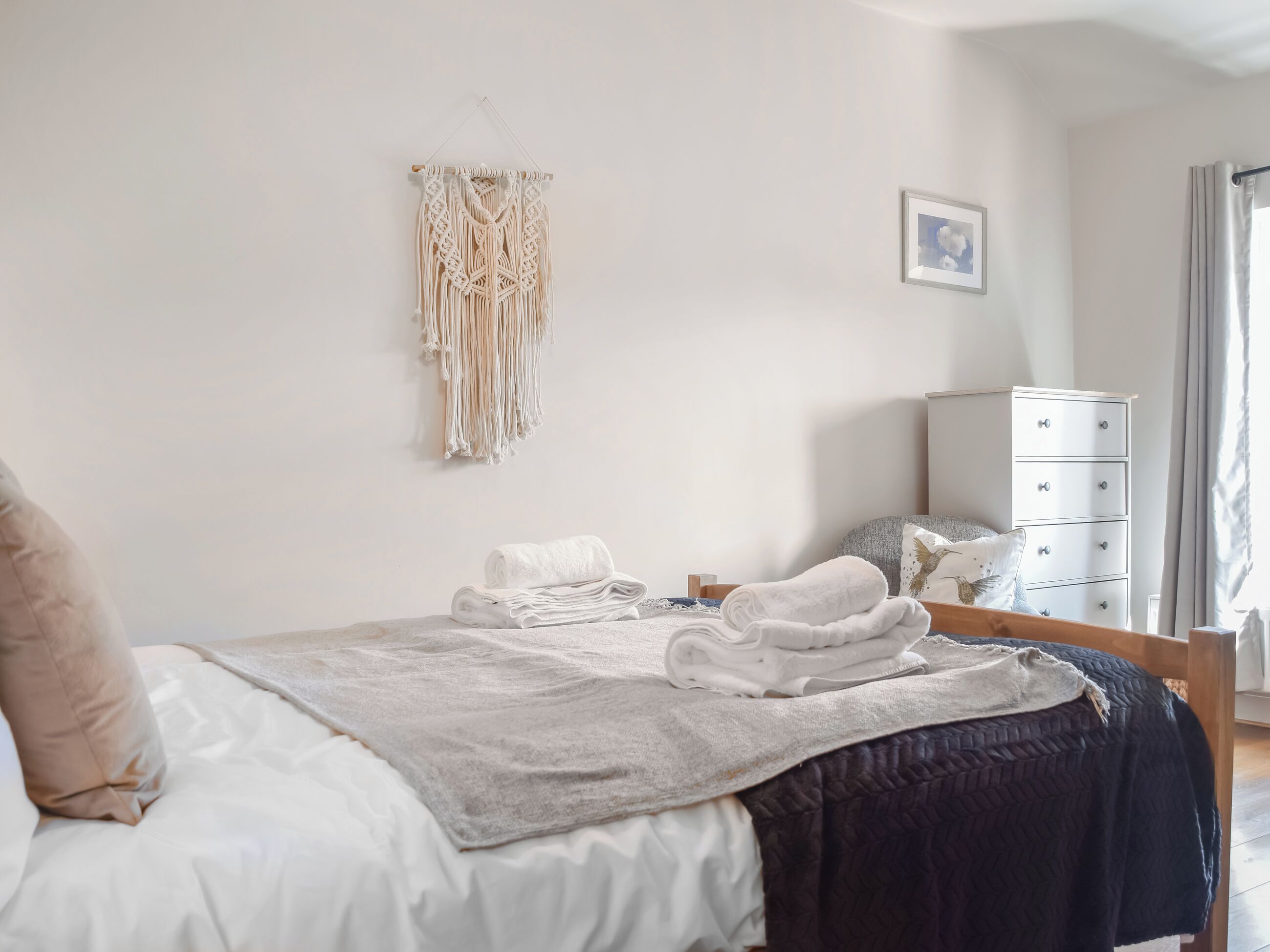 A neatly made bed with a white comforter and a dark blue blanket, a macrame wall hanging, a small bedside table with a lamp, and a white dresser with drawers.