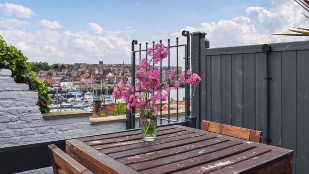A wooden table with a vase of pink flowers, a stone wall with ivy, and a view of a marina and town in the background.