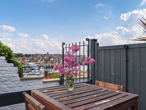 Een houten tafel met een vaas roze bloemen, een stenen muur begroeid met klimop, en een uitzicht op een jachthaven en stad op de achtergrond.