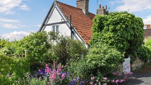 A traditional half-timbered house with a red-tiled roof, surrounded by lush greenery and flowering plants.