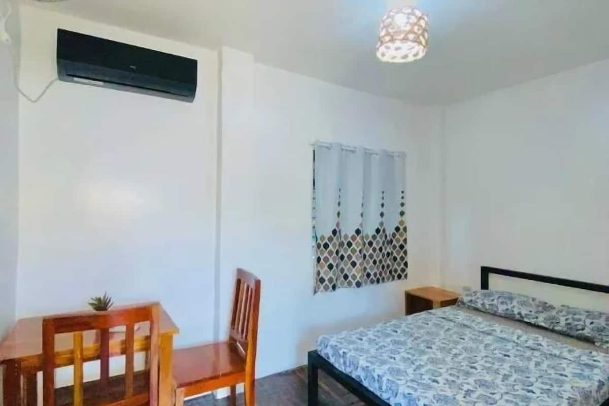 Un dormitorio con cama, escritorio, silla, ventilador de techo y ventana con cortinas estampadas.