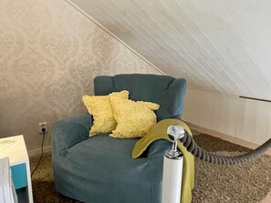 A room with a high ceiling and a sloped ceiling, a teal armchair with two yellow pillows, a small table with a lamp, and a carpeted floor.