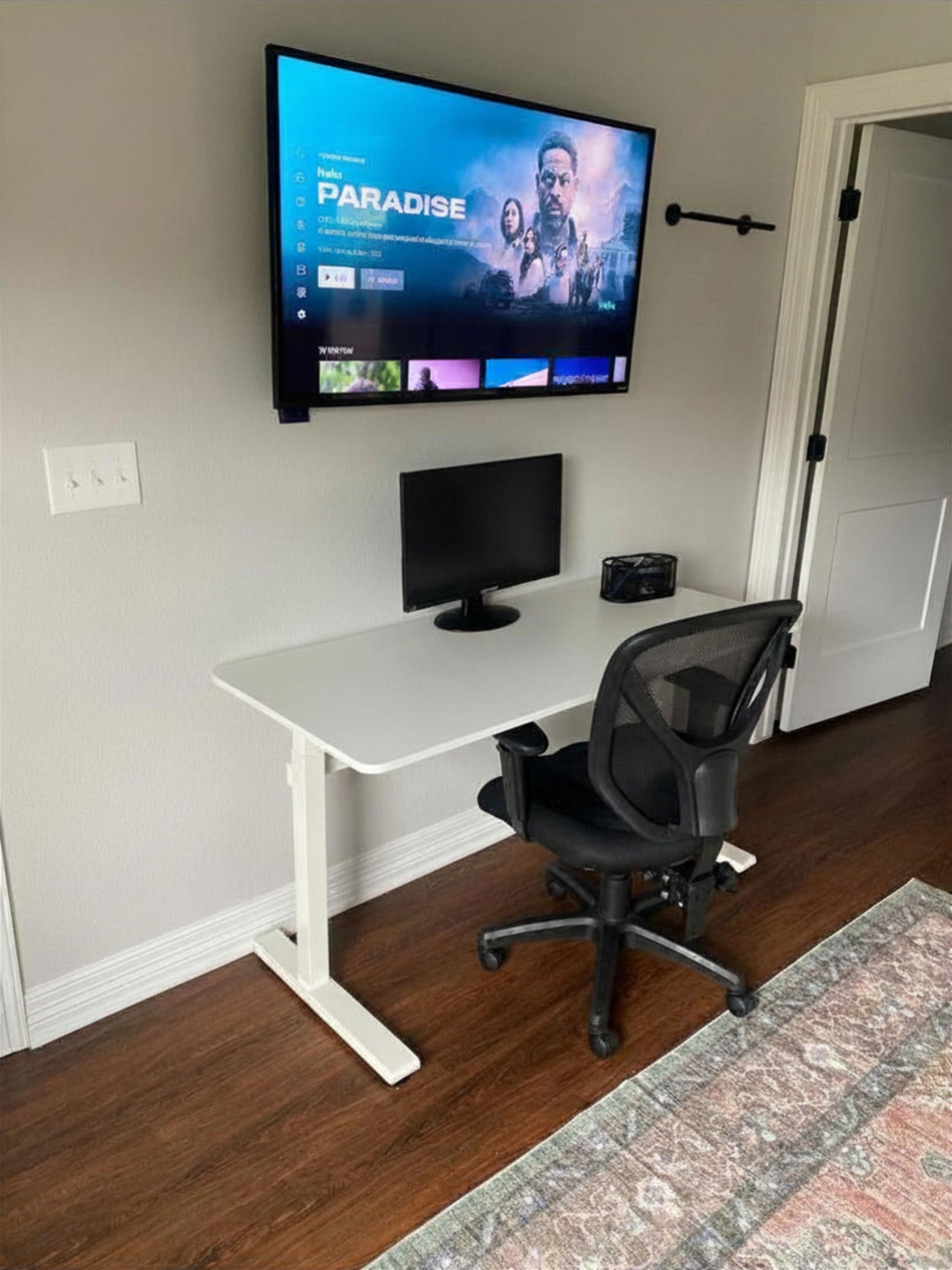 A room with a white desk, a black office chair, a television mounted on the wall, and a door.