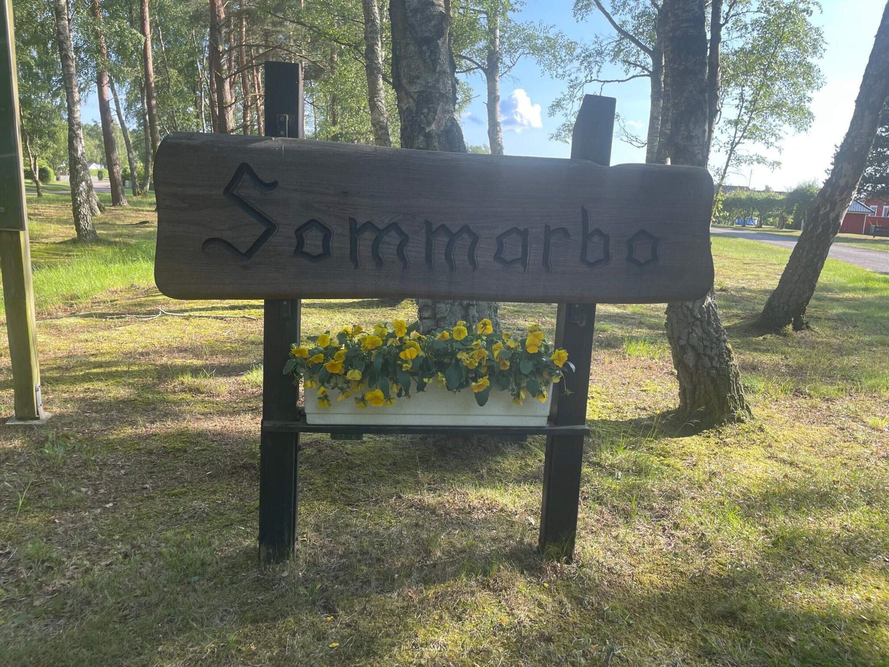Eine hölzerne Bank mit dem eingravierten Namen 'Sommarbo', umgeben von Gras und Bäumen.