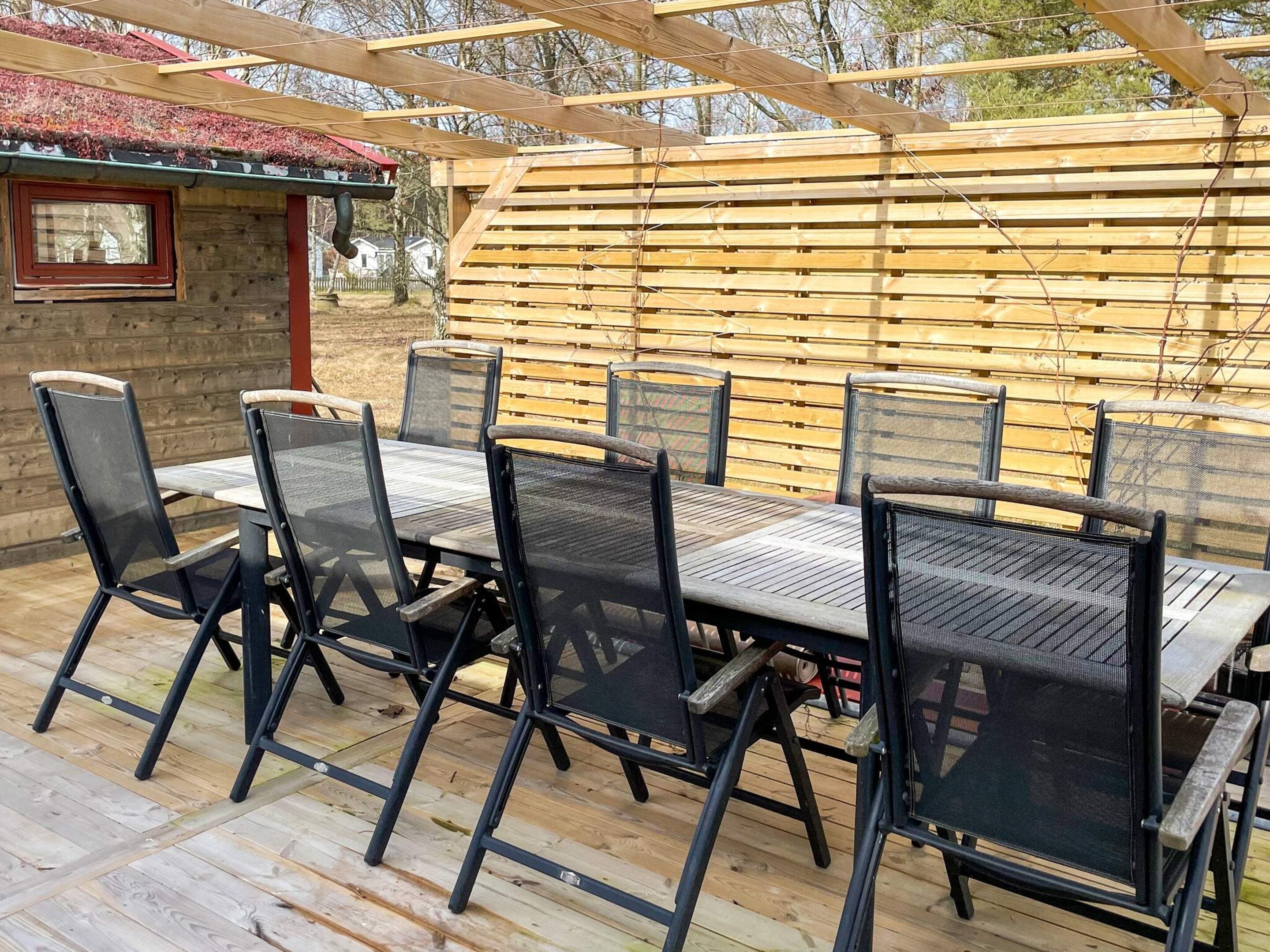 An outdoor patio with a wooden table and chairs, a pergola overhead, and a view of a house in the background.