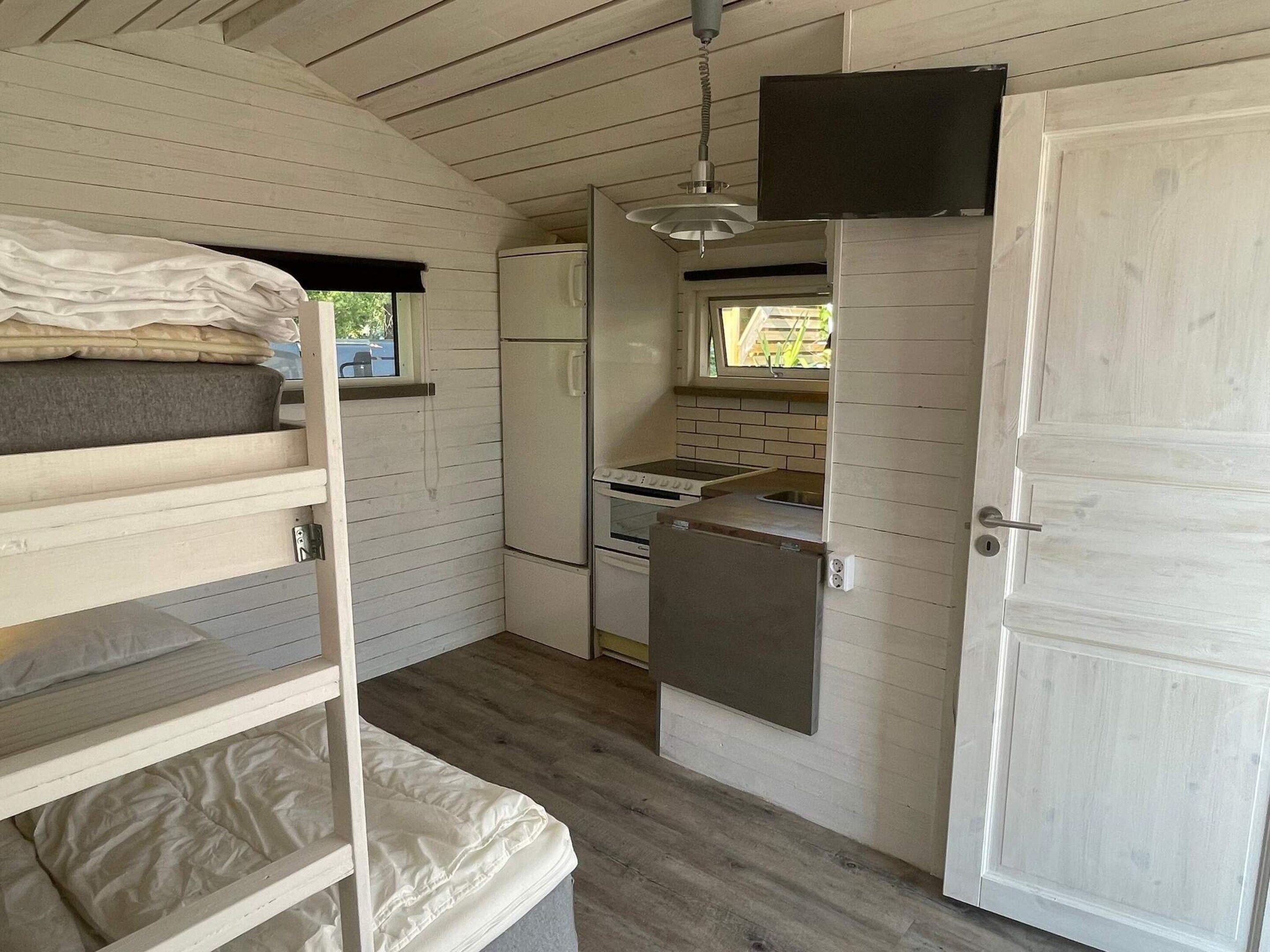 Cocina compacta con refrigerador, estufa y lavamanos, una litera con ropa de cama y un televisor montado en la pared.