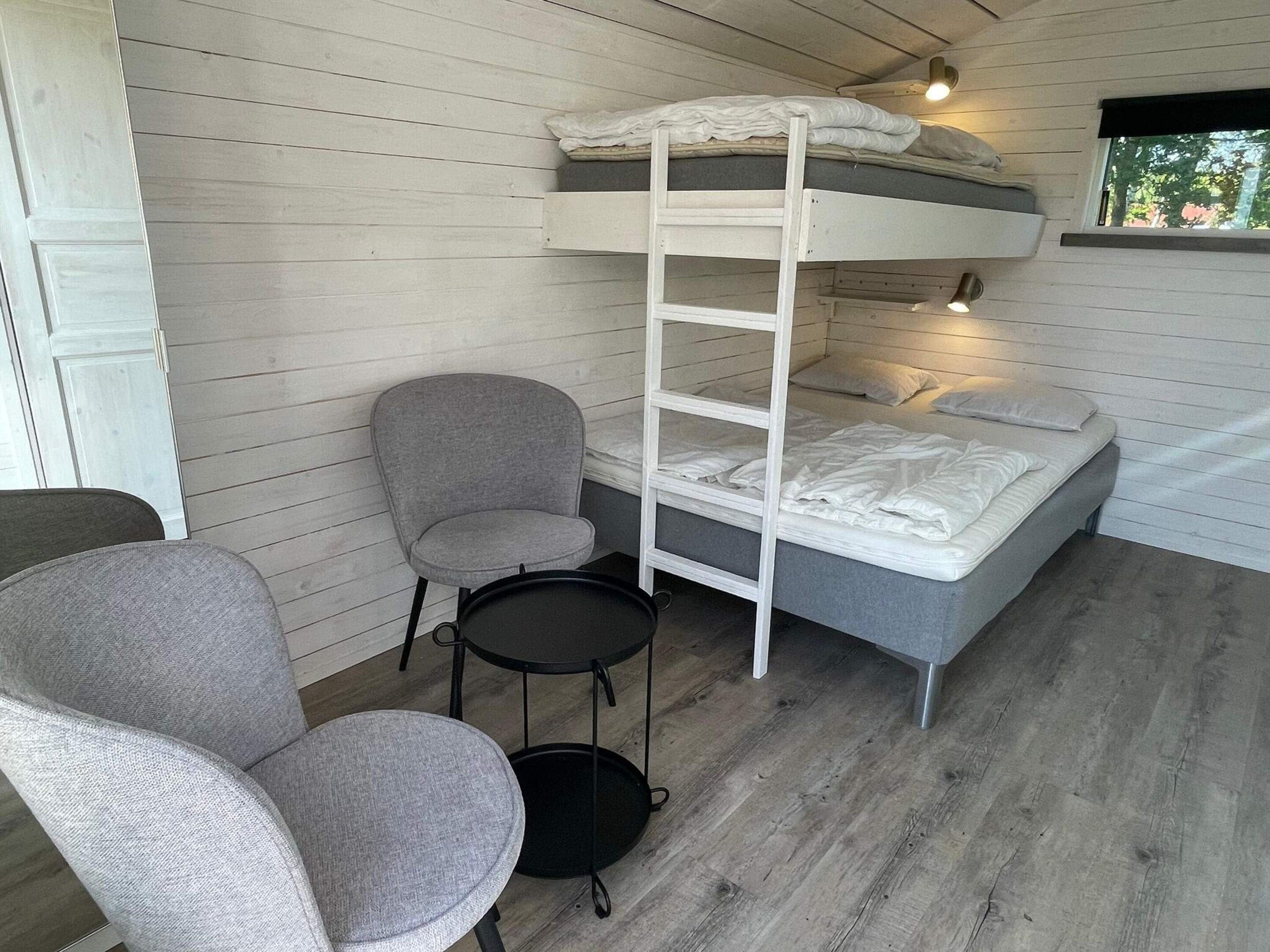 A bunk bed with a ladder, a grey chair, and a small table in a room with wooden walls and a window.