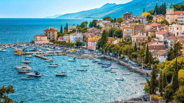 A coastal town with a marina filled with boats, surrounded by colorful buildings and a mountainous backdrop.