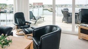 A modern room with a large window overlooking a marina. It includes a black leather chair, a wooden table with a potted plant, and a television on a wooden stand. - 5 Star Holiday Home in Hemmet (Hemmet)