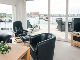 Quarto moderno com uma grande janela com vista para uma marina. Inclui uma poltrona de couro preta, uma mesa de madeira com uma planta em vaso e uma televisão em um suporte de madeira.