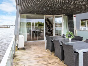 A deck with a table and chairs, a potted plant, and a view of the water and buildings in the background.