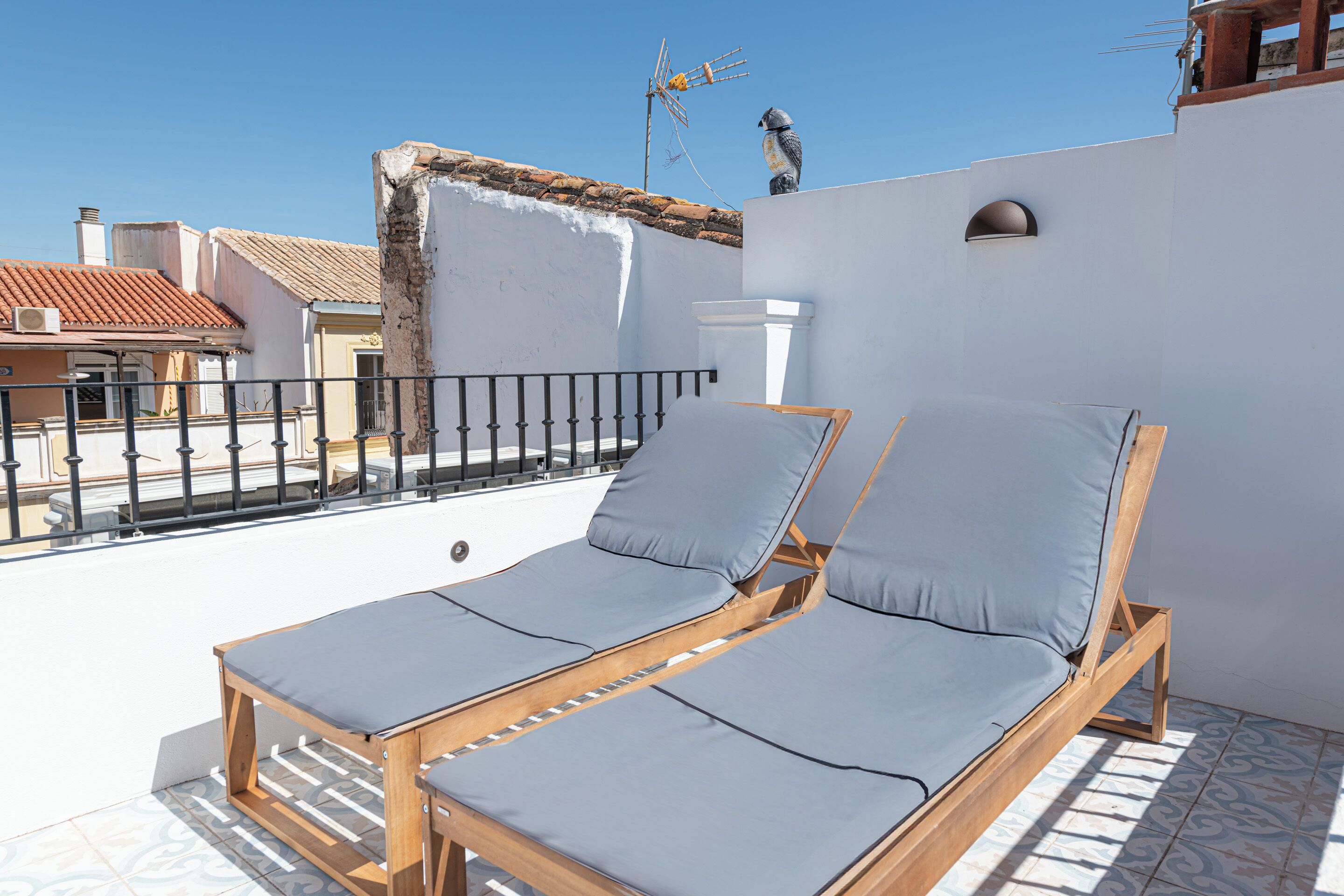 A balcony with two wooden lounge chairs with grey cushions, a white wall, and a tiled floor.