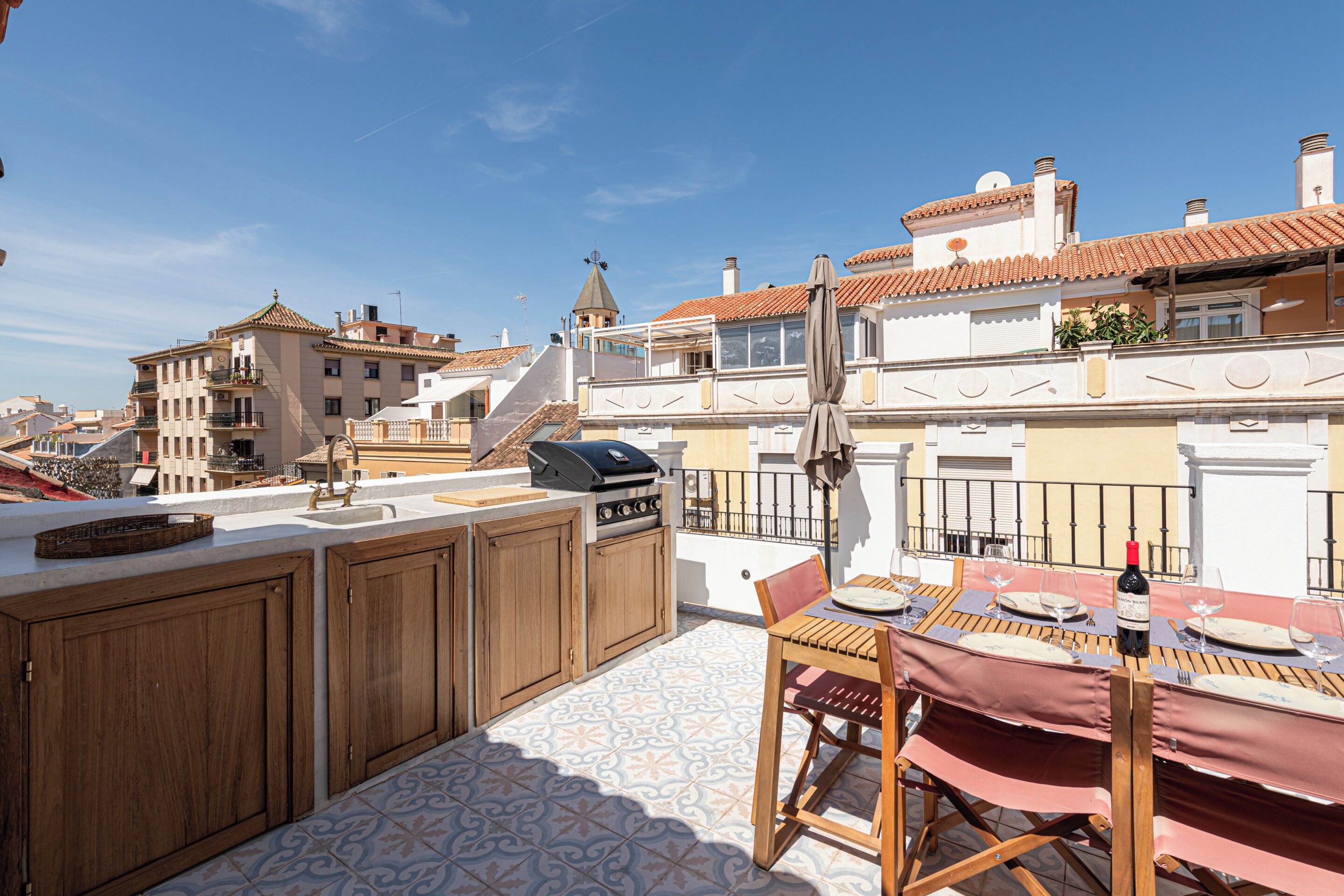A rooftop terrace with a dining table set for two, a grill, and a view of the cityscape.